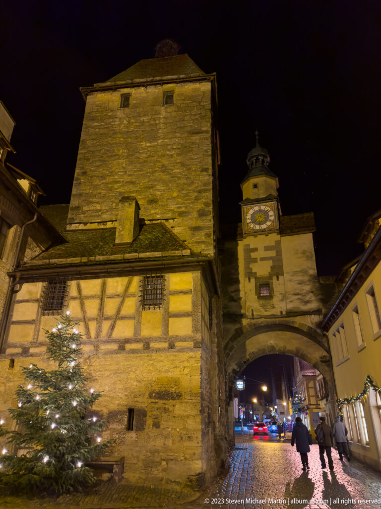 Floodlit medieval gate with massive stone tower, half-timbered lower structure, arched passage, and pedestrians.