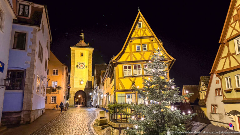 Clocked medieval gate tower beyond a curving cobblestone street, half-timbered house, and decorated Christmas tree.