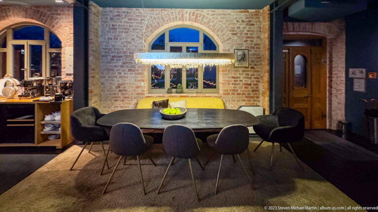 Dining table with six chairs centered beneath a brick arch and rounded window in a modern lodging interior.