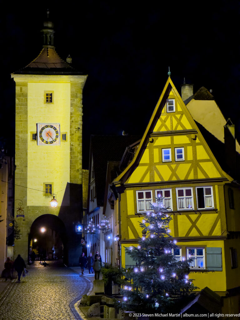 Illuminated clock tower and arched gateway beside a yellow half-timbered house and decorated Christmas tree.