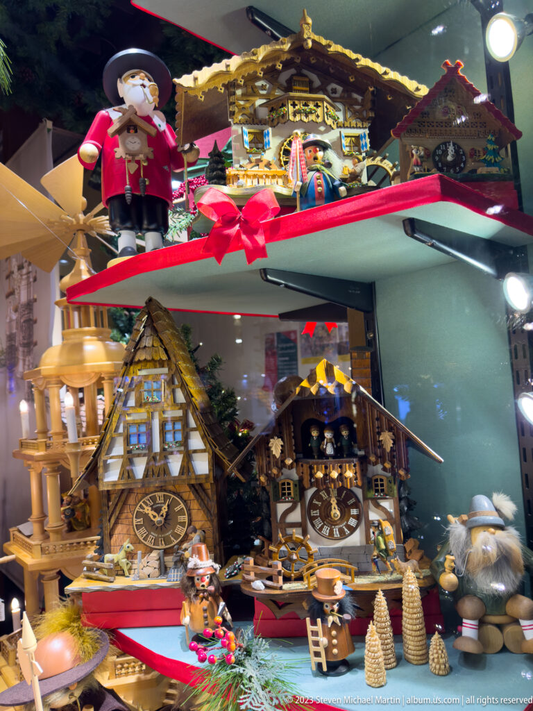 Shop window filled with wooden Christmas pyramids, miniature house clocks, carved figures, and small decorative trees.