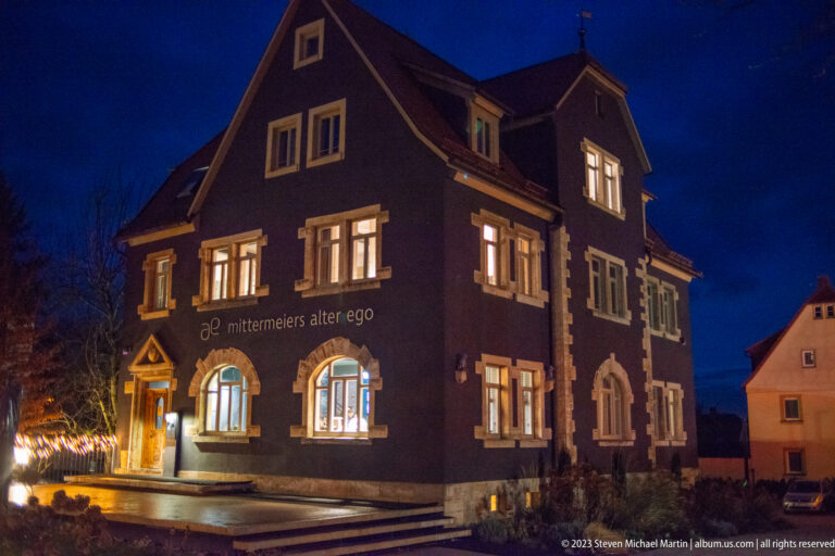 Dark gabled building marked “mittermeiers alter ego,” with lit arched windows and a glowing star decoration outside.