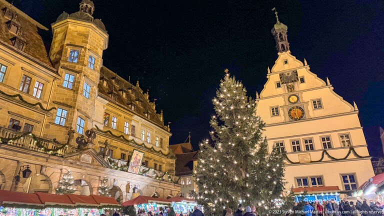 Large lit Christmas tree in Rothenburg’s Marktplatz between the Rathaus and gabled Ratstrinkstube, with market stalls below.