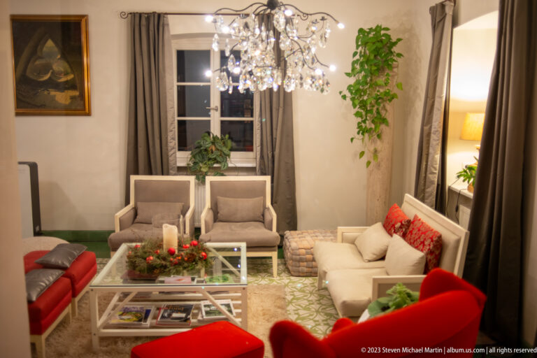 Modern lounge with crystal chandelier, pale seating, red chairs, candles, plants, and a glass coffee table.