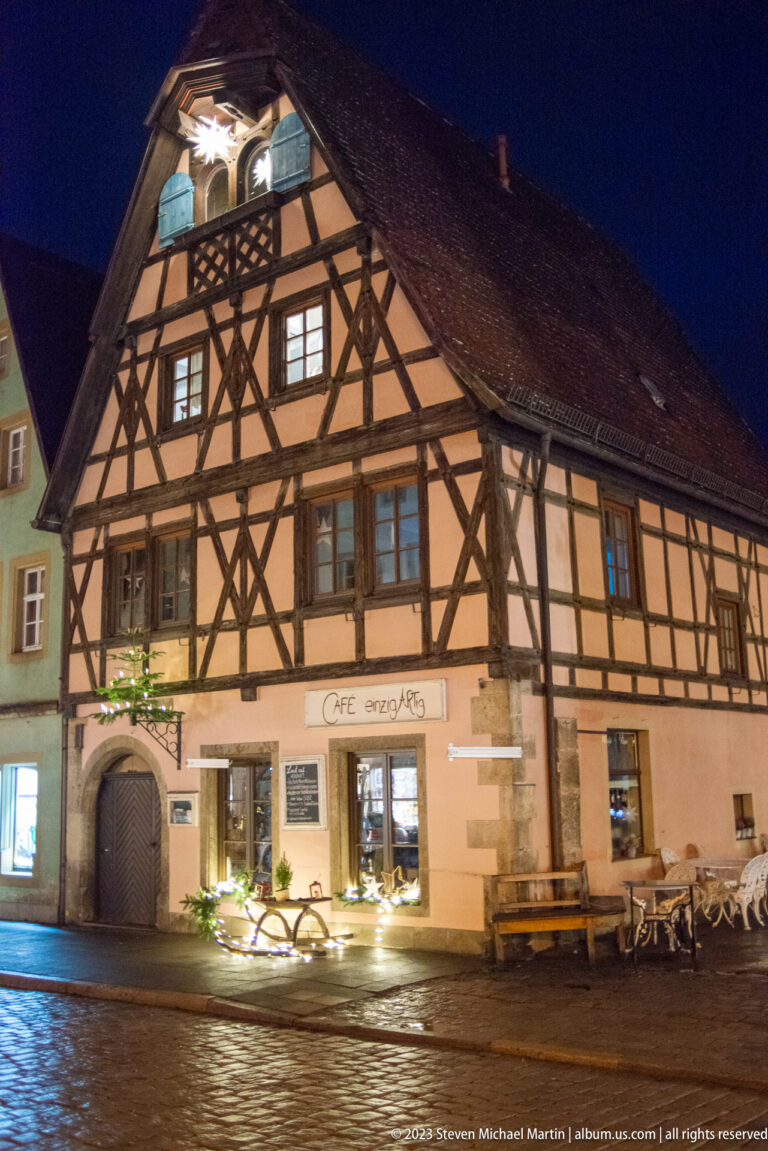 Broad half-timbered building with café windows, benches, and bicycles along a wet street at night.