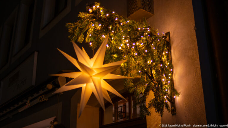 Large glowing many-pointed Christmas star hanging beneath evergreen garland and small white lights on a dark façade.