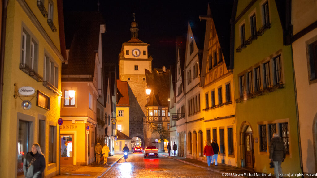 Night street of colorful historic houses leading toward a clocked medieval tower and arched gateway.