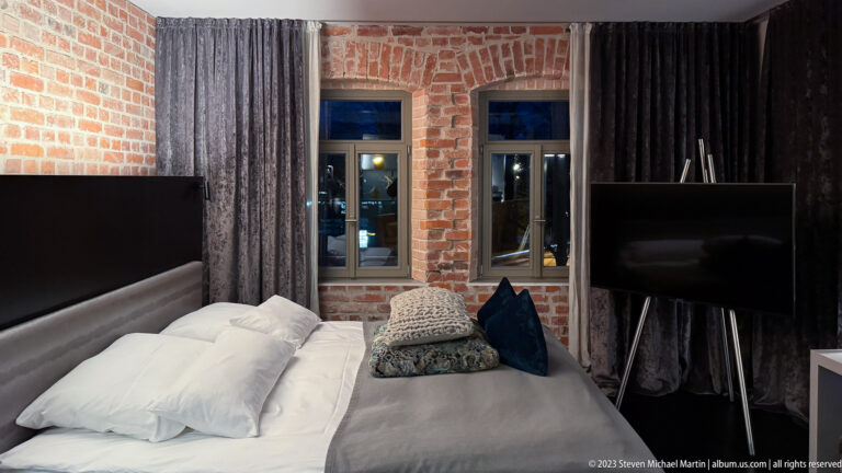 Modern bed beneath tall windows and exposed brick, with heavy curtains and a freestanding television nearby.