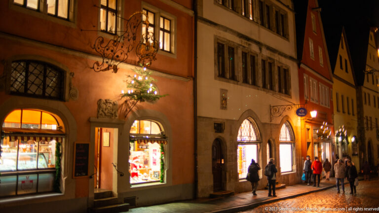Historic shopping street with illuminated windows, projecting signs, Christmas greenery, and pedestrians at night.
