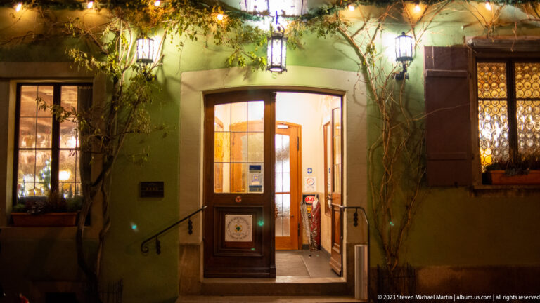 Warmly lit doorway in a green façade with evergreen garland, vines, lanterns, stone steps, and leaded-glass windows.