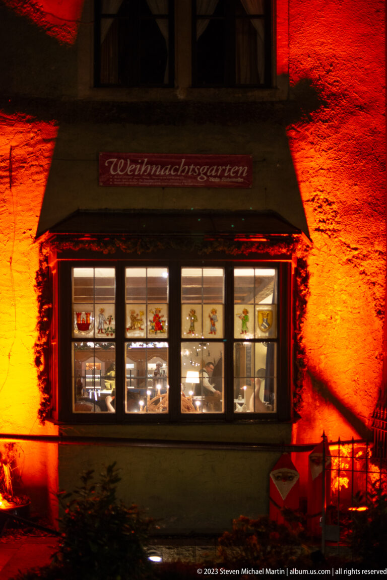 Window beneath a “Weihnachtsgarten” sign, with decorated panes and the surrounding wall lit red-orange.