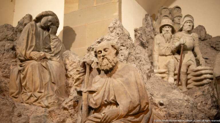 Monochrome museum tableau with a bearded seated figure and armed historical figures posed against a rocky backdrop.