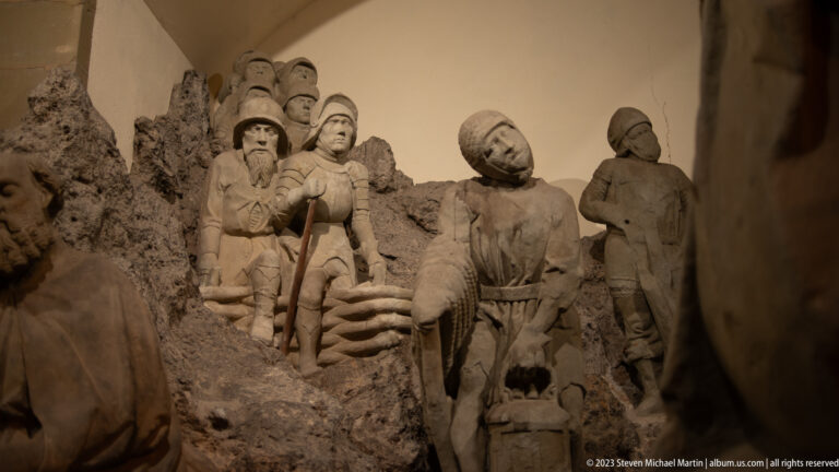 Monochrome museum tableau with a restrained central figure, standing historical figures, and a rocky staged background.