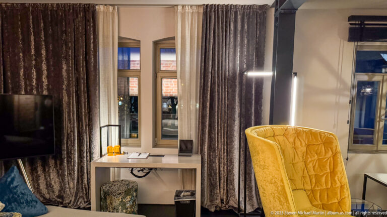 Guest-room desk between curtained windows beside a tall yellow wing chair and vertical task lights.