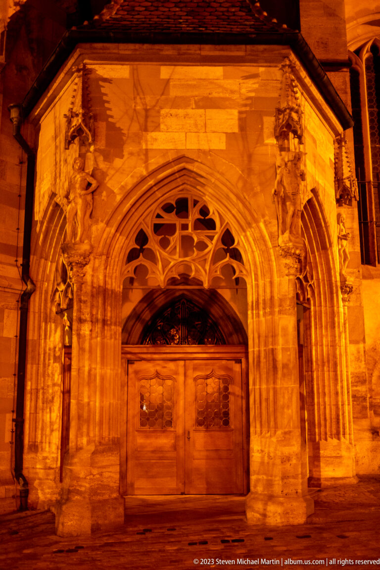Floodlit Gothic stone church portal with pointed arch, carved tracery, sculpted figures, and double wooden doors.