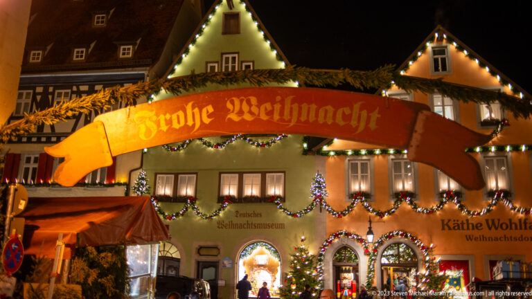 Red “Frohe Weihnacht” arch before the illuminated Käthe Wohlfahrt and German Christmas Museum façades.