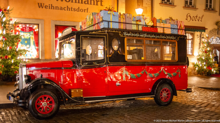 Red and black vintage-style Christmas vehicle with roof gifts and garlands parked outside decorated buildings.