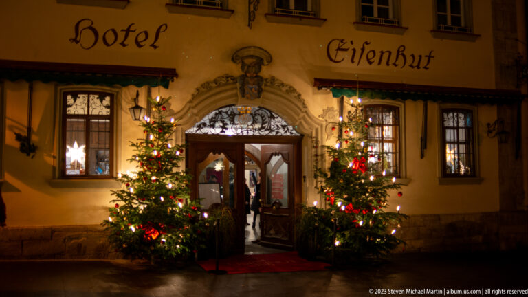 Hotel Eisenhut entrance beneath its painted name, flanked by two lit Christmas trees and carved stonework.