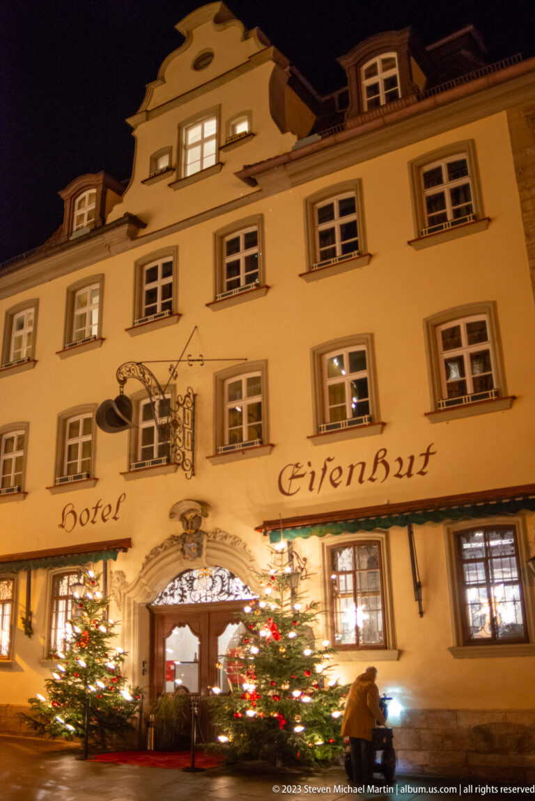 Illuminated Hotel Eisenhut façade with rows of shuttered windows, dormers, and Christmas trees around the entrance.