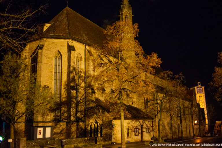 Floodlit Gothic St. James’s Church with tall windows, buttresses, steep roof, tower, and bare winter trees.