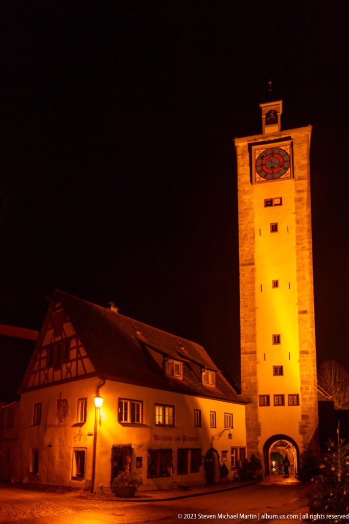 Floodlit rectangular medieval gate tower with arched passage beside a long historic building at night.