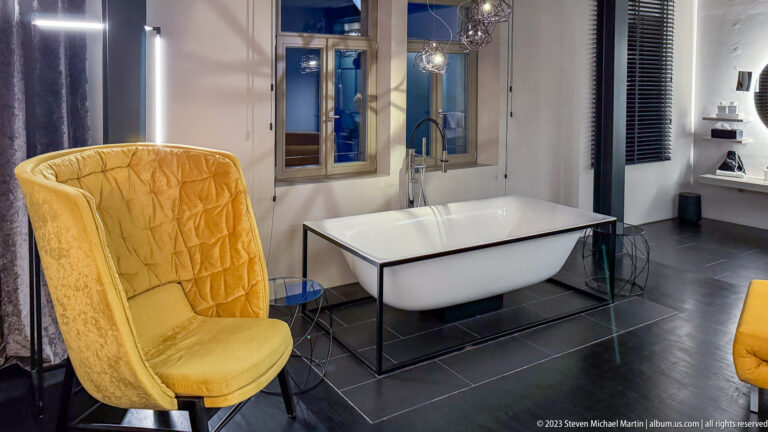 Freestanding white bathtub on dark tile beside a yellow wing chair and tall windows in a modern bathroom.