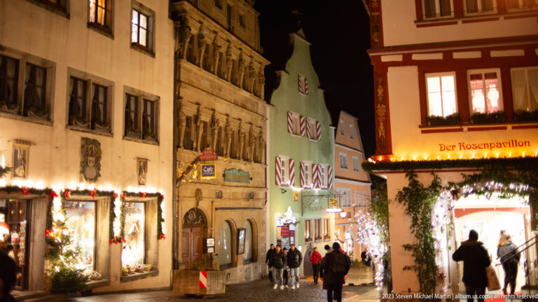 Historic lane with pedestrians, decorated shopfronts, garlands, and the illuminated “Der Rosenpavillon” storefront.