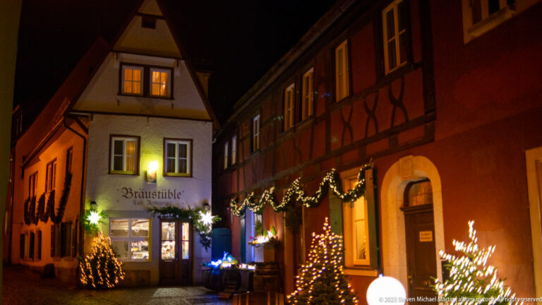 Bräustüble façade at the end of a decorated lane with garlands, small lit trees, and a red timbered building.