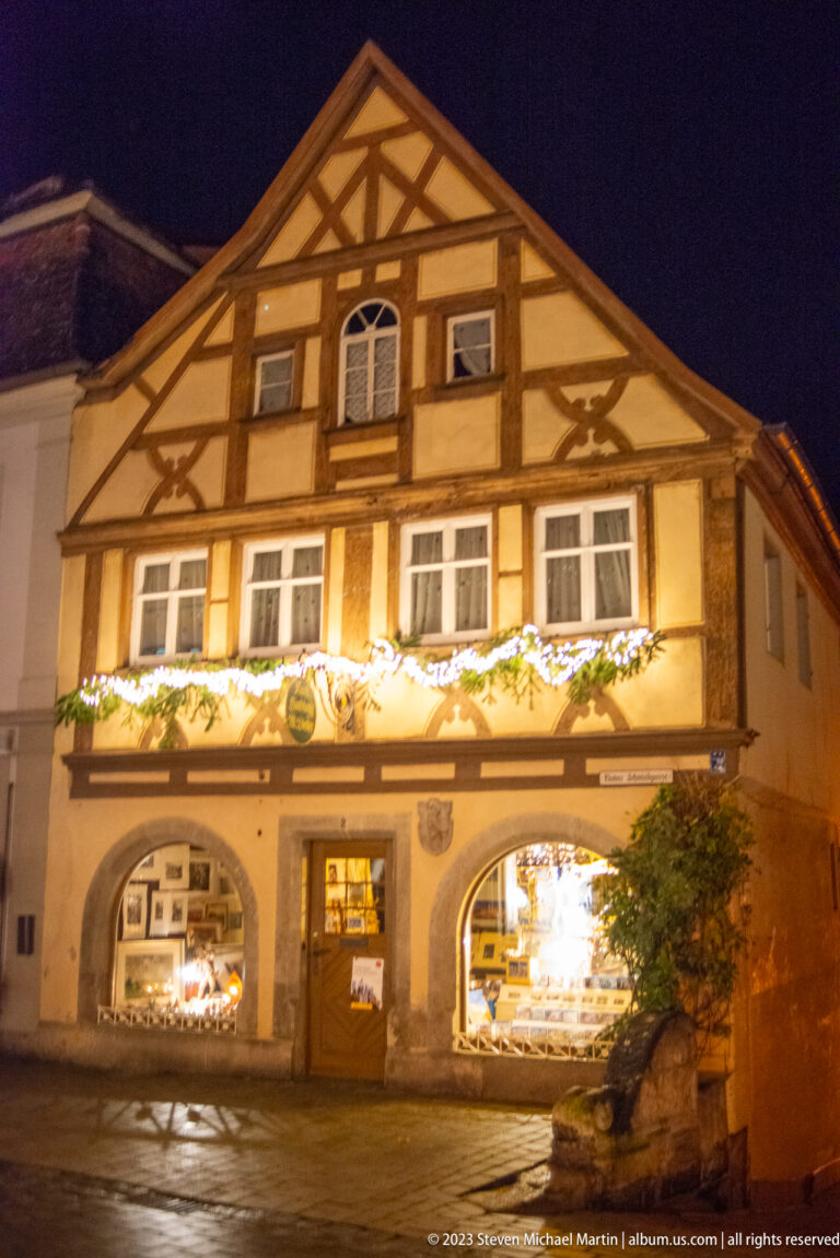 Broad gabled half-timbered building with lit arched shop windows and Christmas garland across the façade.