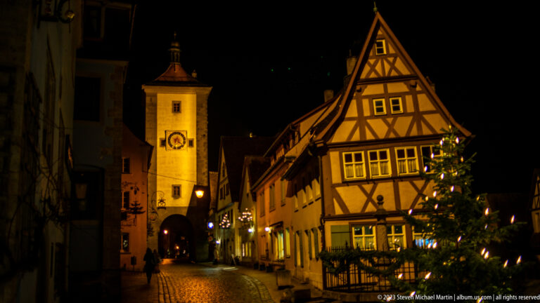 Illuminated Markusturm and Röderbogen beside a steep half-timbered house and decorated Christmas tree.