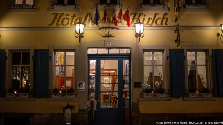 Hotel Hirsch entrance beneath international flags, with blue shutters, wall lanterns, and illuminated doorway.