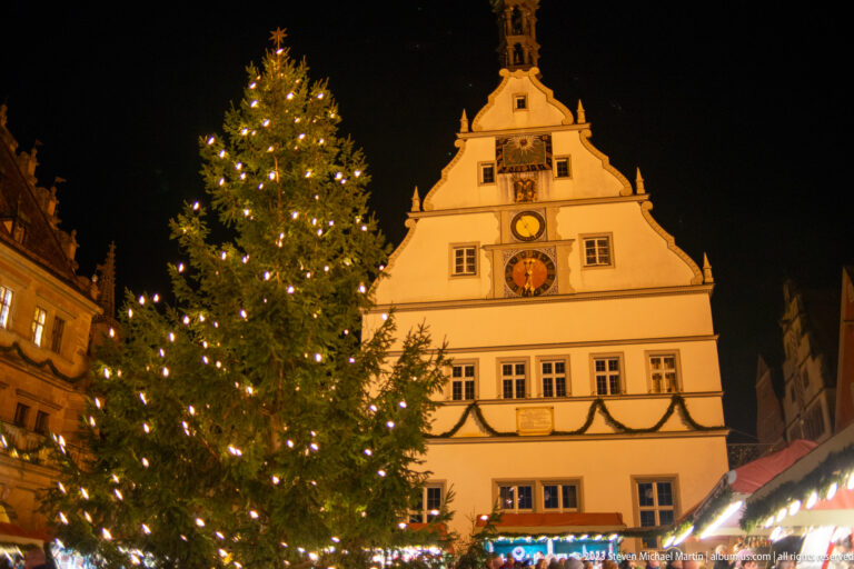 Large lit Christmas tree before the white stepped-gable Ratstrinkstube with illuminated clock faces.