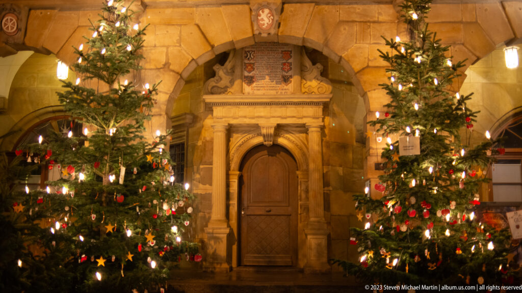 Ornate stone doorway with columns, pediment, heraldic shields, wooden door, and Christmas trees on both sides.