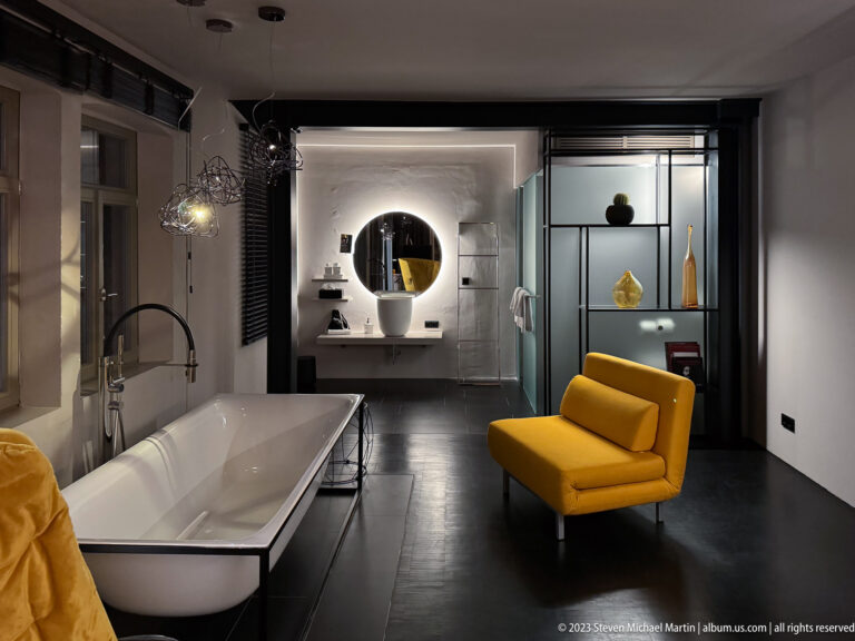 Open-plan hotel bathroom with freestanding tub, yellow lounge chair, glass shelving, and a brightly lit vanity.