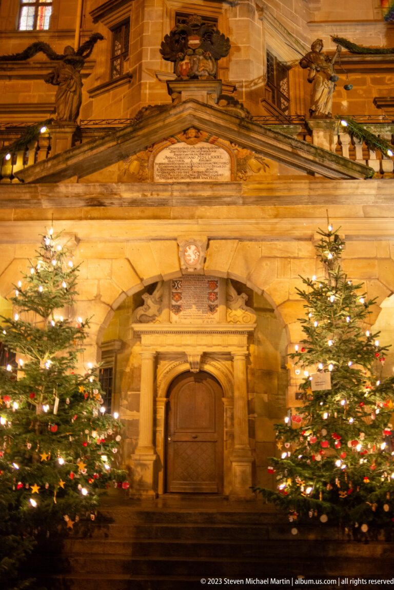 Wide view of an ornate stone civic portal between two lit Christmas trees, with carved reliefs above.