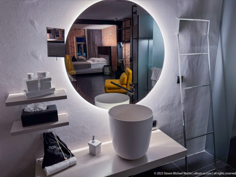 Backlit round mirror above a white vessel sink, reflecting a bed, yellow chair, and exposed brick in the adjoining room.