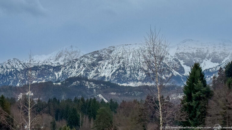 Snow-covered Alpine ridges rise beyond dark evergreen and leafless woodland near Füssen.