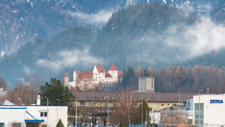 High Castle of Füssen with pale walls and red roofs on a wooded hill above modern buildings.