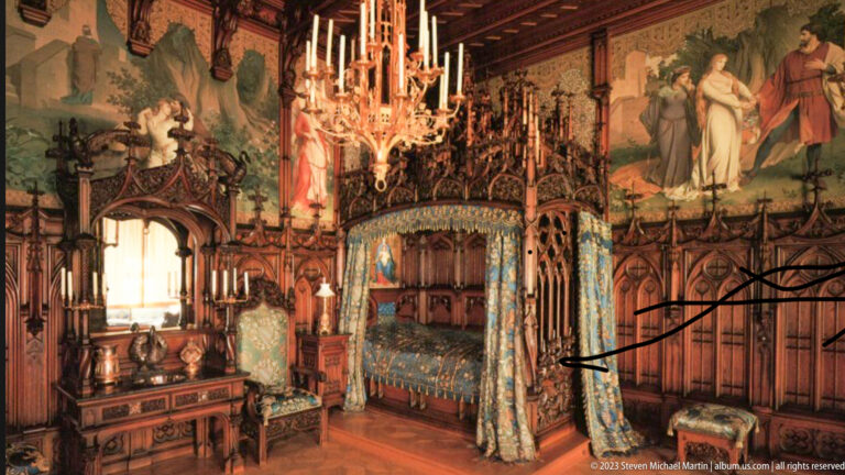 Canopied bed with blue textiles, carved woodwork, chandelier, and large wall murals in Hohenschwangau Castle.
