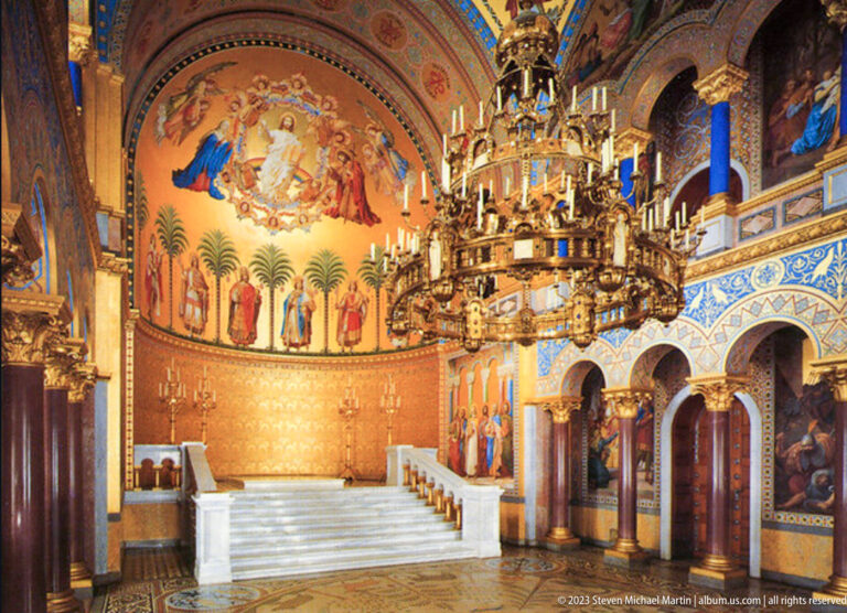 Blue-and-gold ceremonial hall with white steps, painted arcades, murals, columns, and a large chandelier.
