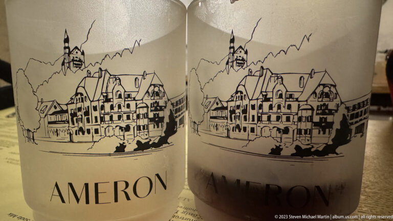Two white Ameron mugs printed with a black line drawing of a castle, village roofs, and mountains.