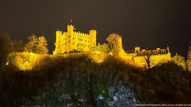 Hohenschwangau Castle illuminated in warm gold light above a dark wooded ridge at night.