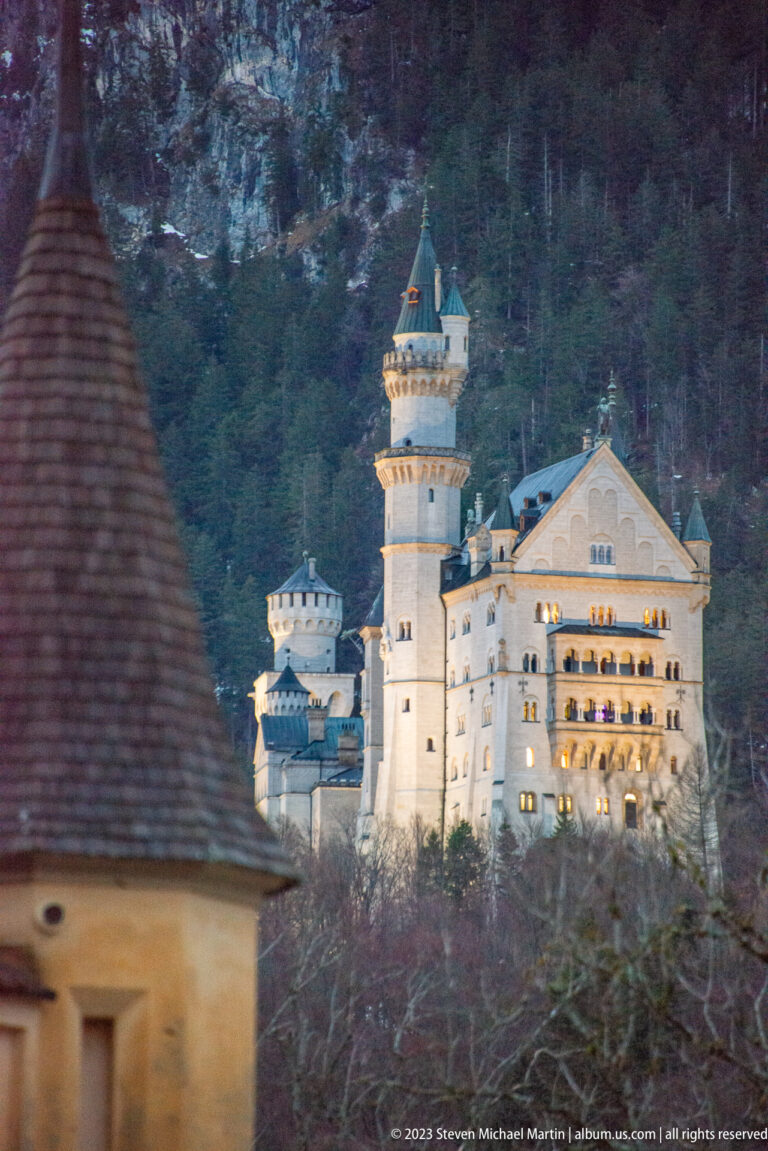 Neuschwanstein Castle at dusk with illuminated windows, pale towers, and a dark forested mountainside behind.