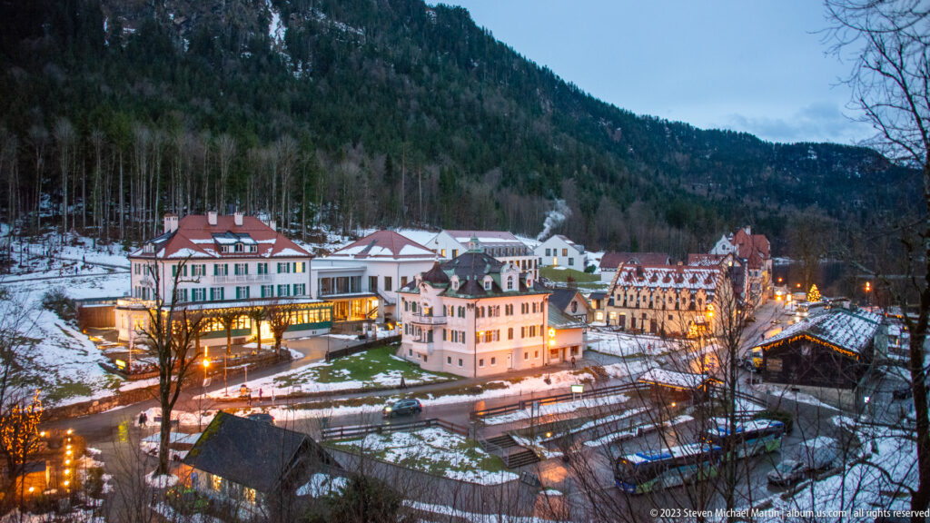 Snowy Hohenschwangau visitor district at dusk with lit hotels and buildings beneath a forested mountain.