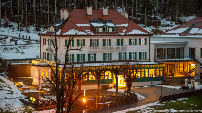 Large pale hotel with green shutters, red roof, and brightly lit lower dining level in snowy Hohenschwangau.