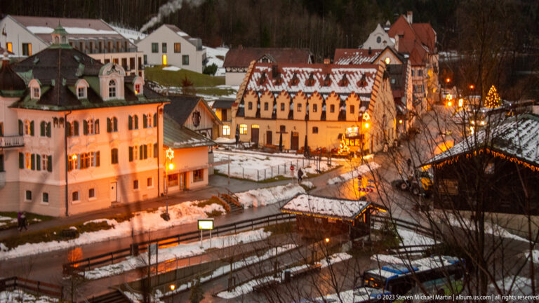 Snow-covered Hohenschwangau buildings and paths lit by warm lamps at night beneath a dark hillside.