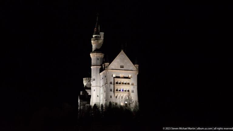 Neuschwanstein Castle illuminated against a black night sky, with its tall tower and pale palace walls visible.
