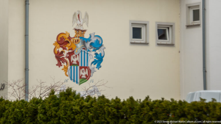 Colorful painted coat of arms with shield, helmet, crest, and red-blue mantling on a cream wall.