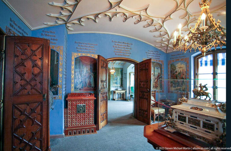 Blue Hohenschwangau room with carved doors, painted inscriptions, decorative stove, chandelier, and museum barriers.