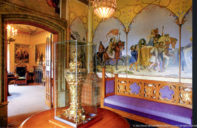 Silver-colored decorative object in a glass case before a large mural and purple-upholstered carved bench.